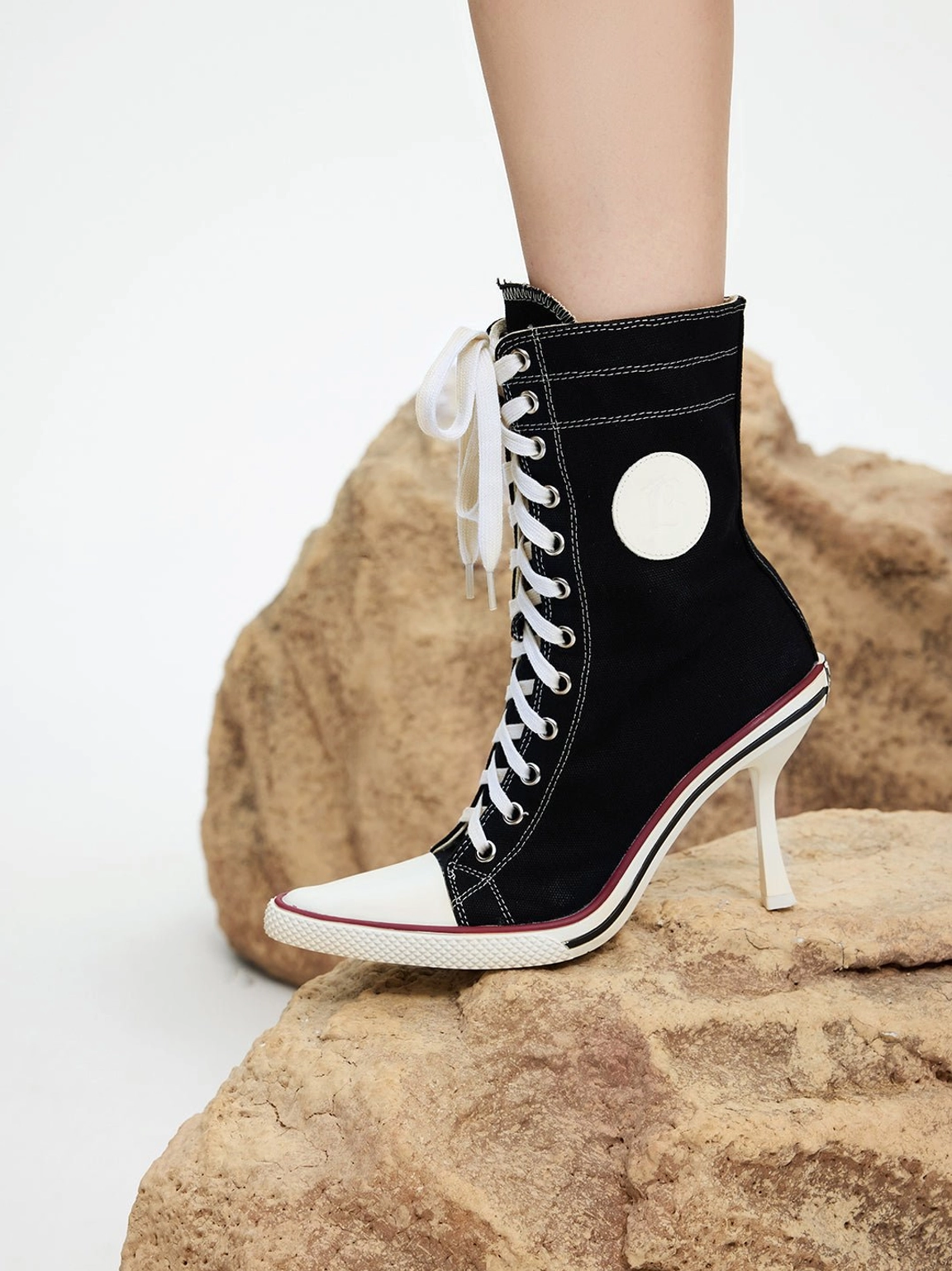 Close-up detailed view of canvas stiletto boot on model ankle with lace-up closure oversized laces round ankle patch and slim heel clearly visible on brown rock prop, macro product photography