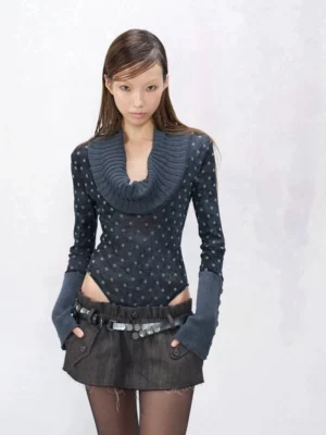 Front view of blue grey polka dot cowl neck knit bodysuit with dramatic ribbed neckline and extended cuffs styled with a mini denim skirt and sheer tights