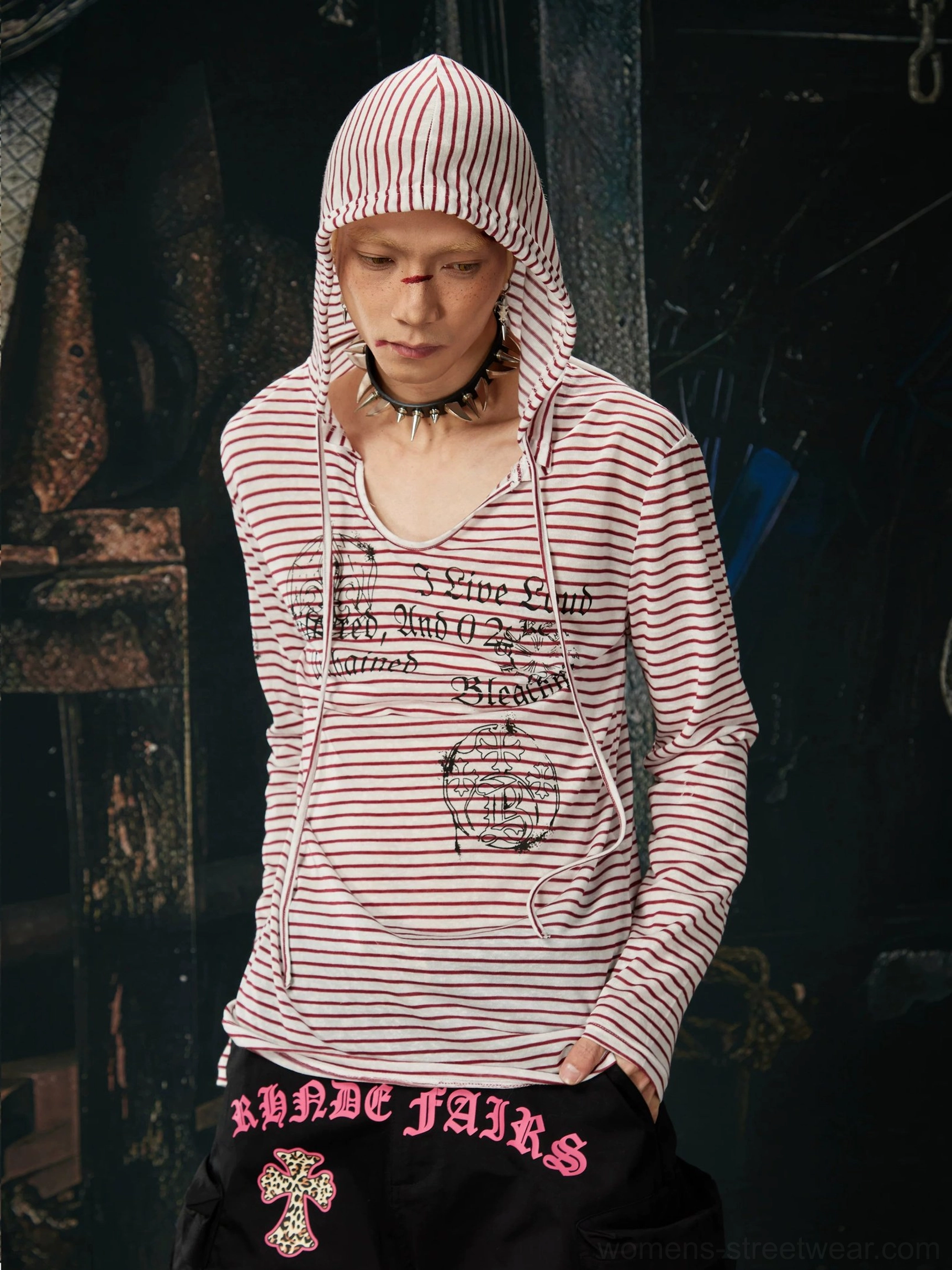 Front close-up of a striped gothic hooded long sleeve streetwear top with drawstring V-neck, script chest graphics, and black cargo pants in an industrial workshop