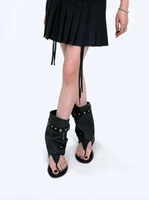 Model wearing black studded buckle thong ankle boot sandals with a black pleated mini skirt
