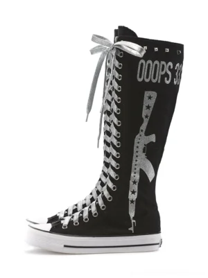 Black studded graphic canvas knee-high punk boots shown from a three-quarter front view with silver laces, rivet collar trim, printed lettering, star motifs, white toe caps, and flat rubber soles