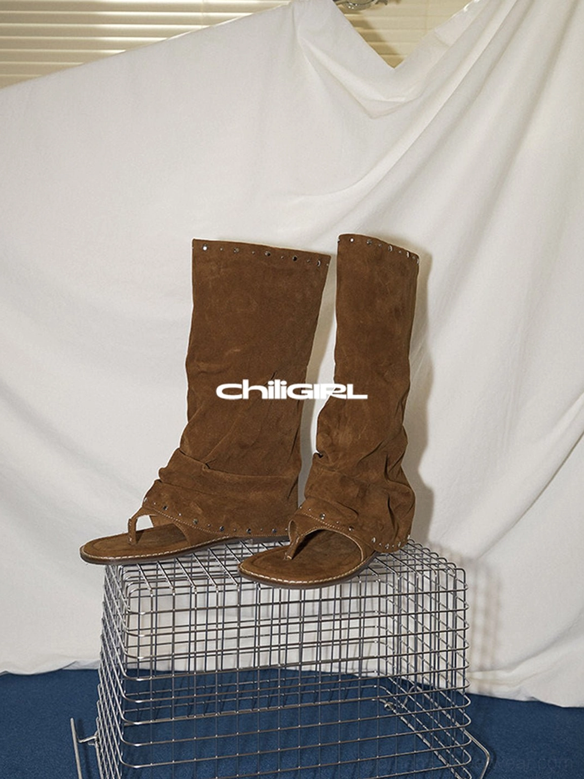 Brown studded suede thong sandal boots displayed on a metal grid stand, showing the tall ruched shaft and flat sole
