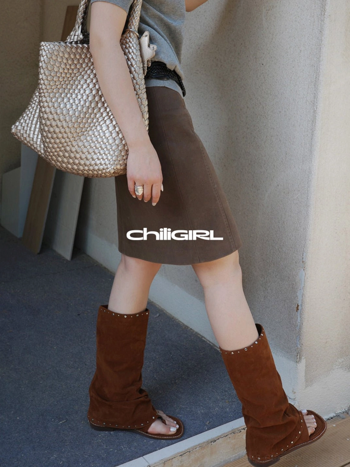 Side view of brown studded suede open-toe tall sandal boots styled with a brown midi skirt and woven tote bag
