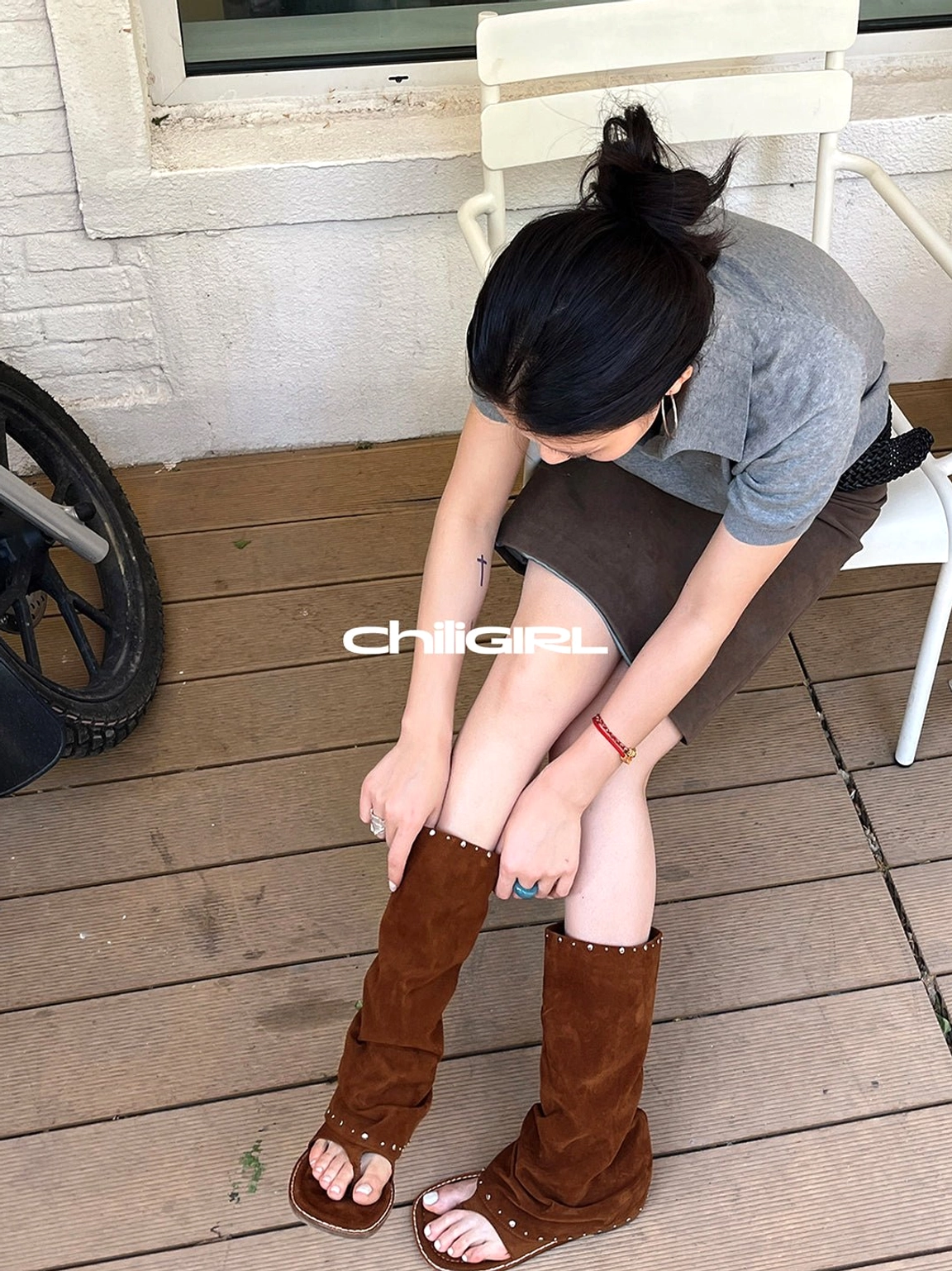 Overhead view of a model putting on brown studded suede thong sandal boots with silver rivet edging and a ruched shaft