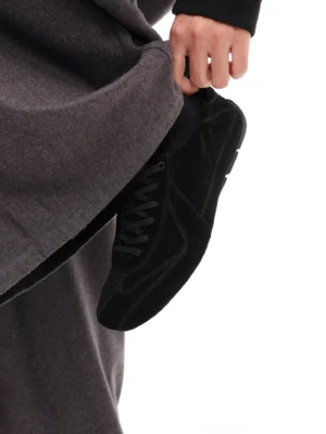 Black suede minimalist lace-up sneaker held by hand with grey trousers, showing a sleek low-top German trainer-inspired streetwear silhouette