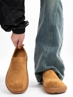 Tan suede backless mule clogs worn with baggy blue jeans and a black windbreaker, showing the closed round toe, chunky low platform sole, and minimalist streetwear slip-on design