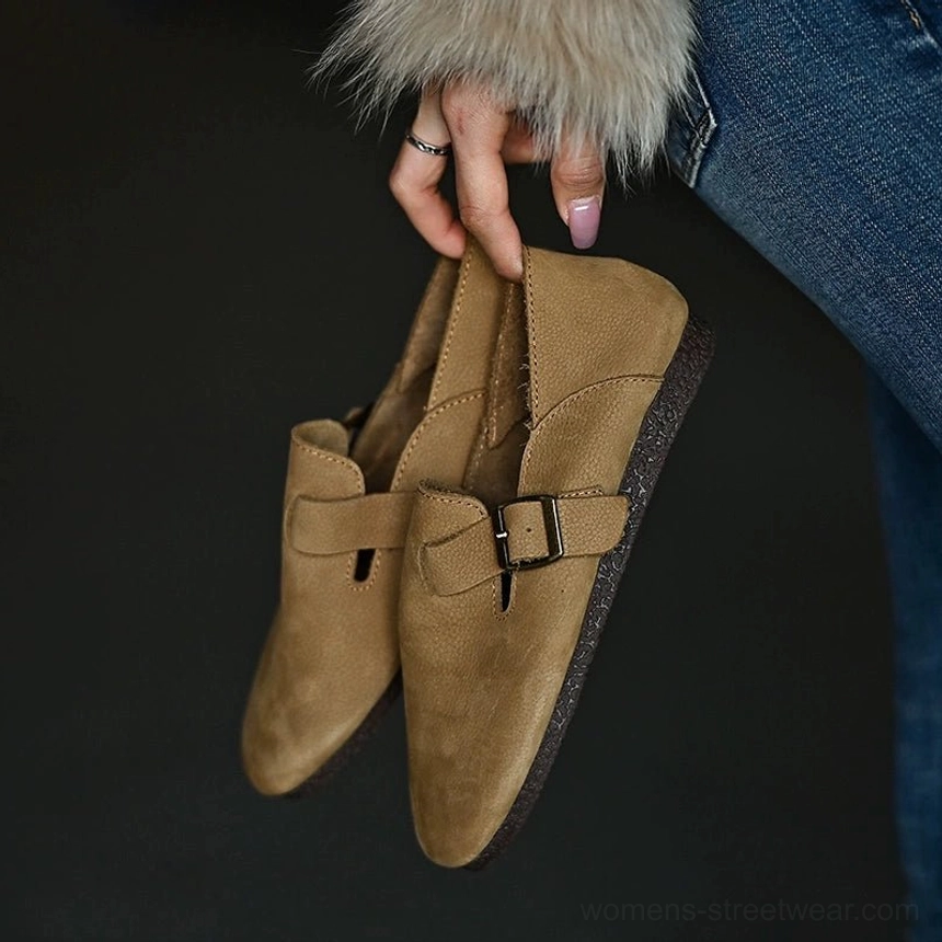 Close-up of a pair of tan leather buckle loafers held in hand, clearly showing the sole, buckle hardware, and collapsible heel