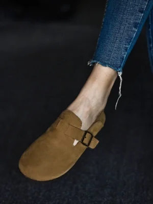 Side close-up of a tan leather slip-on loafer with dark adjustable buckle, low flat sole, and soft textured finish