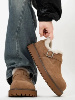 Tan suede shearling buckle platform clogs lifted in a low side view, showing the plush fleece collar, adjustable silver buckle, and chunky lug rubber sole