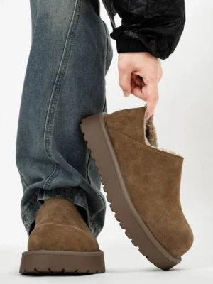 Tan suede shearling-lined platform slip-on clogs worn with blue jeans and black streetwear outerwear, showing the chunky rubber sole and cozy faux fur lining
