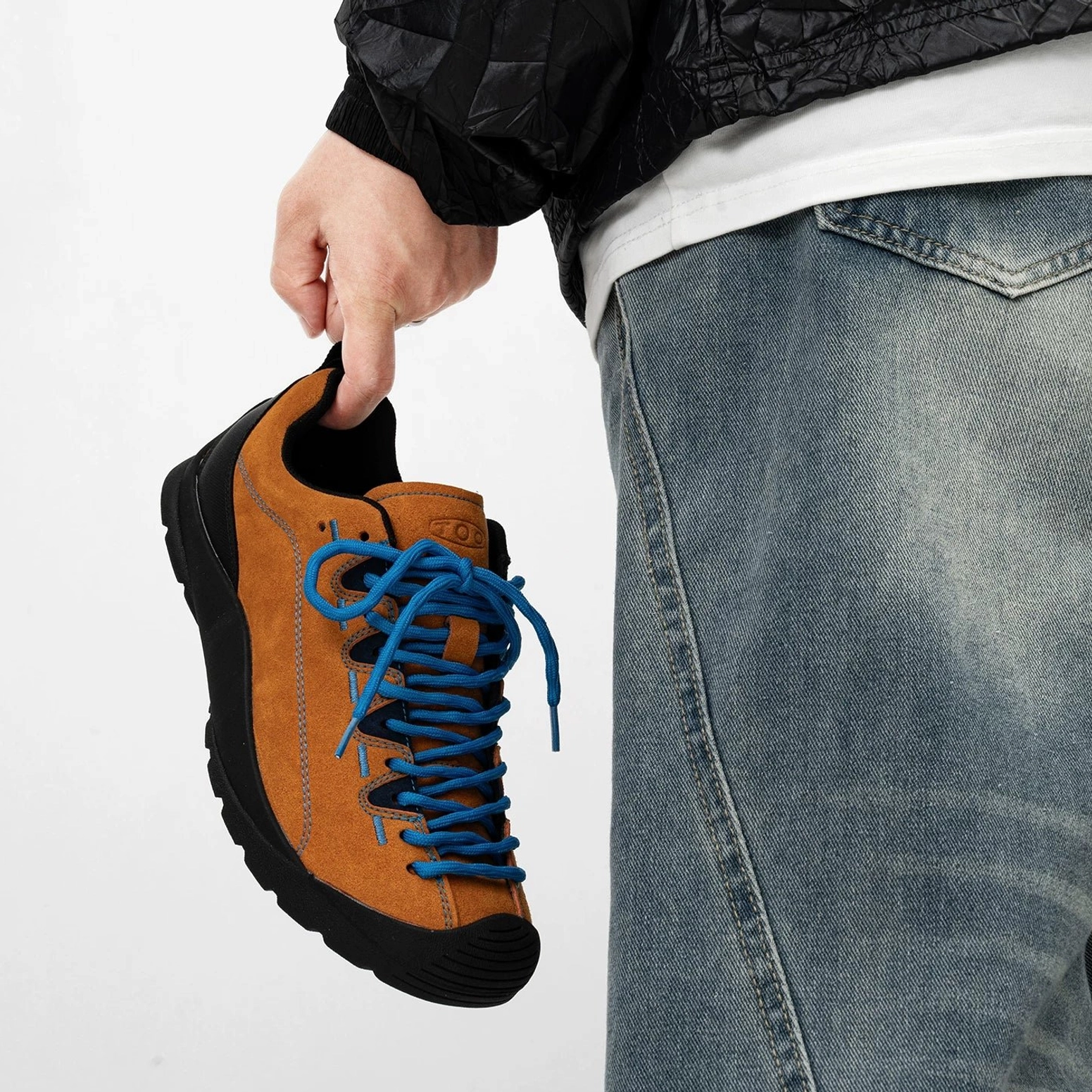 Close hand-held side view of tan suede trail sneakers with blue rope laces, extended eyelets, black toe bumper and rubber outsole