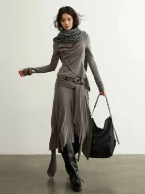 Full body front three quarter view of a taupe asymmetric draped two piece streetwear set with long sleeve top, waterfall wrap skirt, scarf, belt, ankle boots, and tote bag