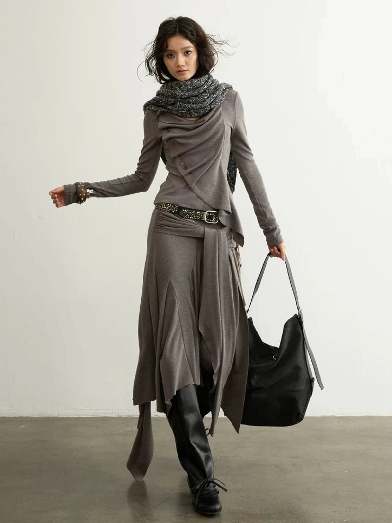 Full body front three quarter view of a taupe asymmetric draped two piece streetwear set with long sleeve top, waterfall wrap skirt, scarf, belt, ankle boots, and tote bag