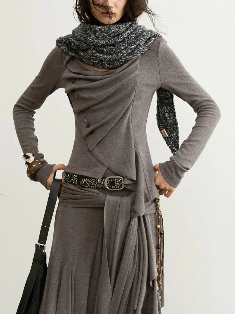 Close up front view of taupe asymmetric draped long sleeve knit top with layered folds, small front buttons, sculptural hem, and studded belt styling