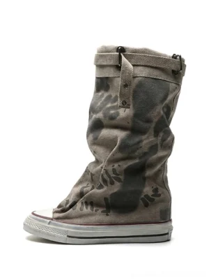 Taupe graffiti canvas slouchy platform tall boots with faded camouflage print, rubber toe caps, lace-up fronts, and buckle collar straps on a white background