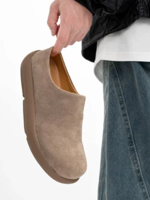 Taupe suede backless mule clogs shown from an overhead side angle with layered casual streetwear clothing, highlighting the smooth matte upper and chunky low-profile sole