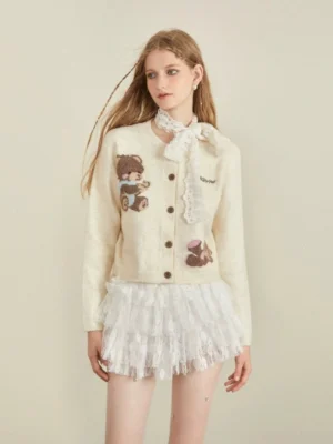 Front view of a cream beige cropped knit cardigan featuring two teddy bear jacquard prints, crew neckline, button front, and Korean streetwear styling