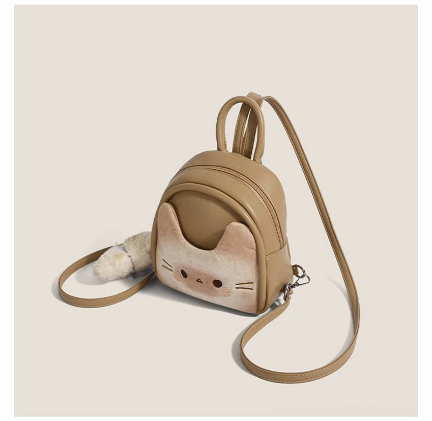 Back left three quarter view of a toast beige cat mini backpack showing the shoulder straps, side zipper hardware, front cat face panel, and detached fluffy puppy pendant
