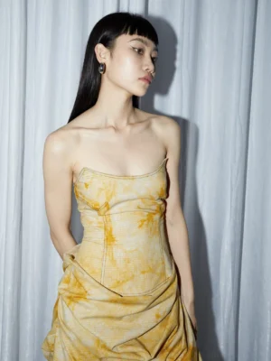 Front three-quarter view of a turmeric yellow ruched strapless tie-dye midi dress with corset-inspired bodice and sculptural draped skirt
