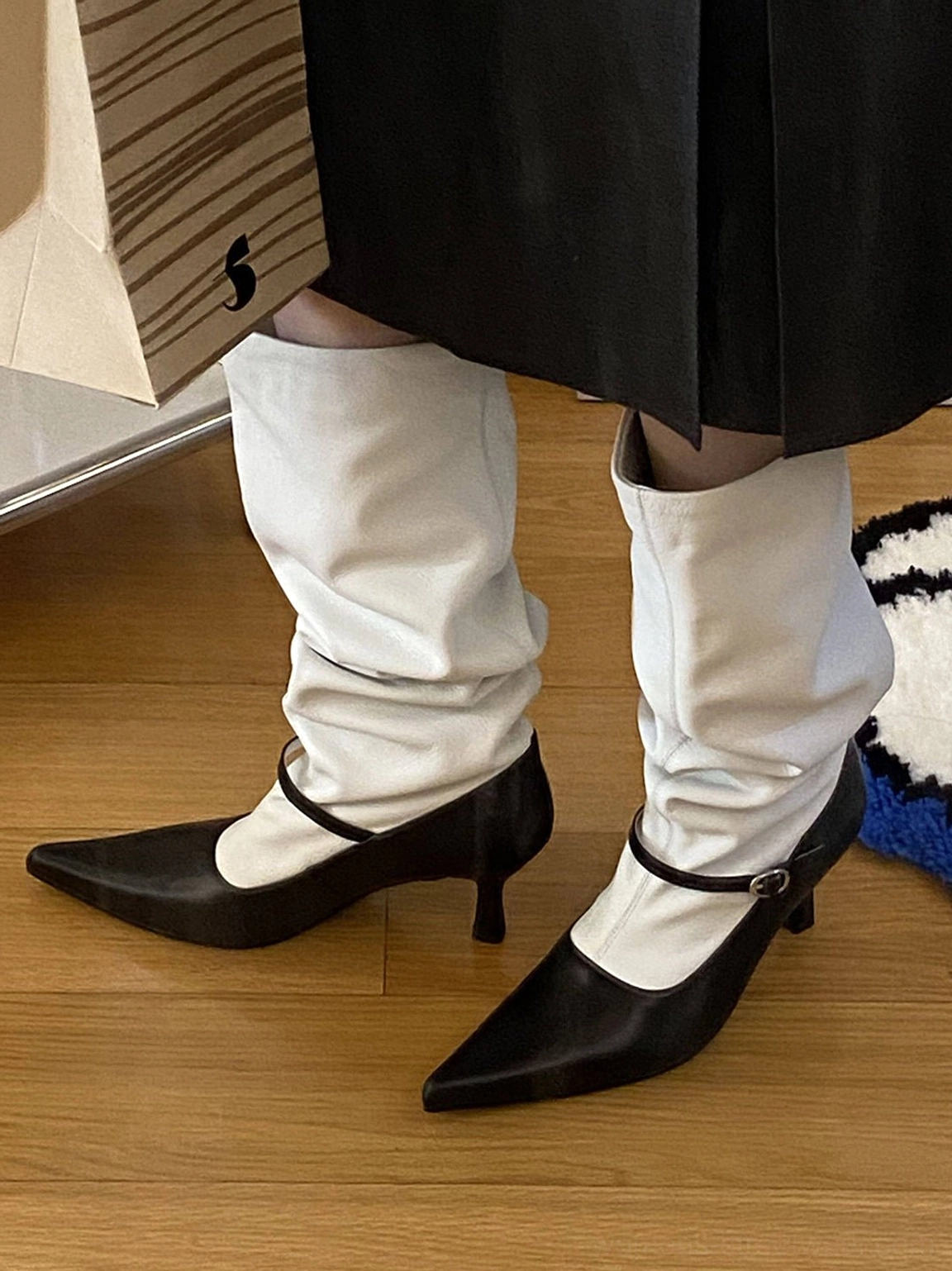 Front three-quarter view of two-tone black and white pointed-toe knee-high boots styled with a black outfit, slouchy shaft, and Mary Jane-inspired ankle straps