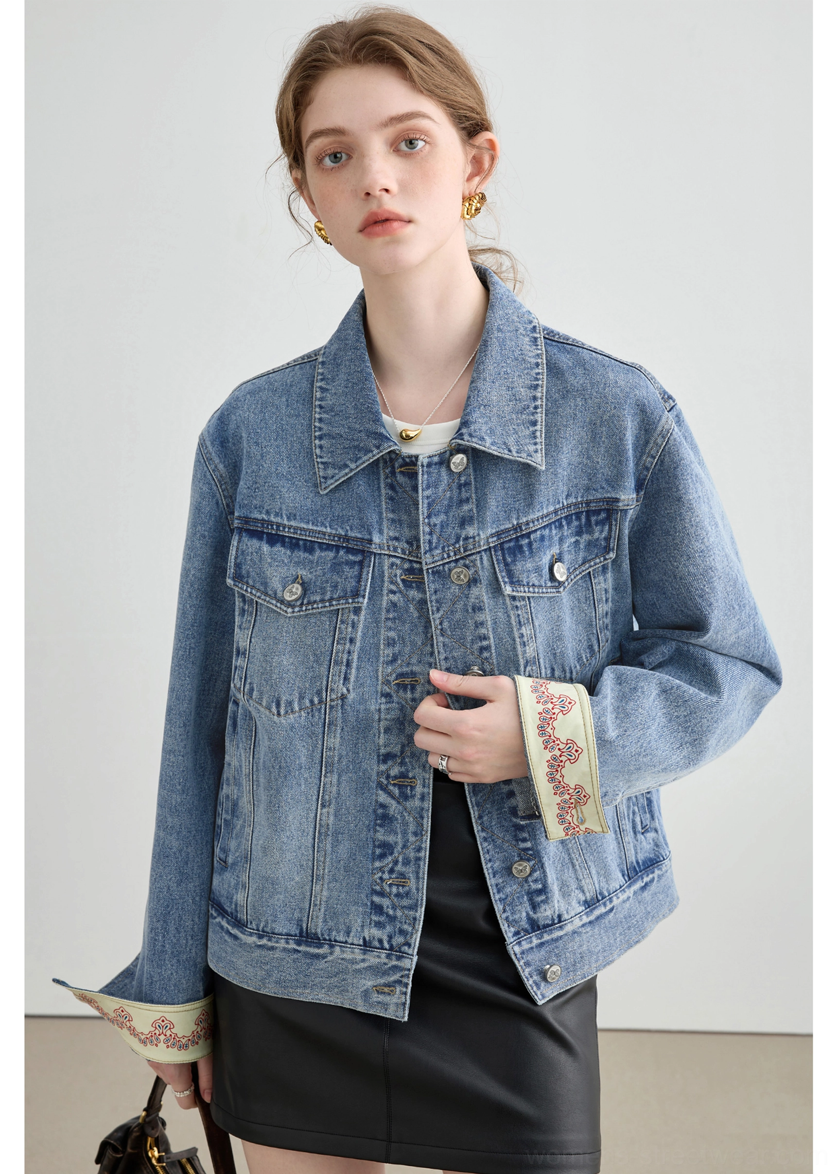Close-up of a female model wearing an unbuttoned light wash denim trucker jacket with patterned embroidered cuffs