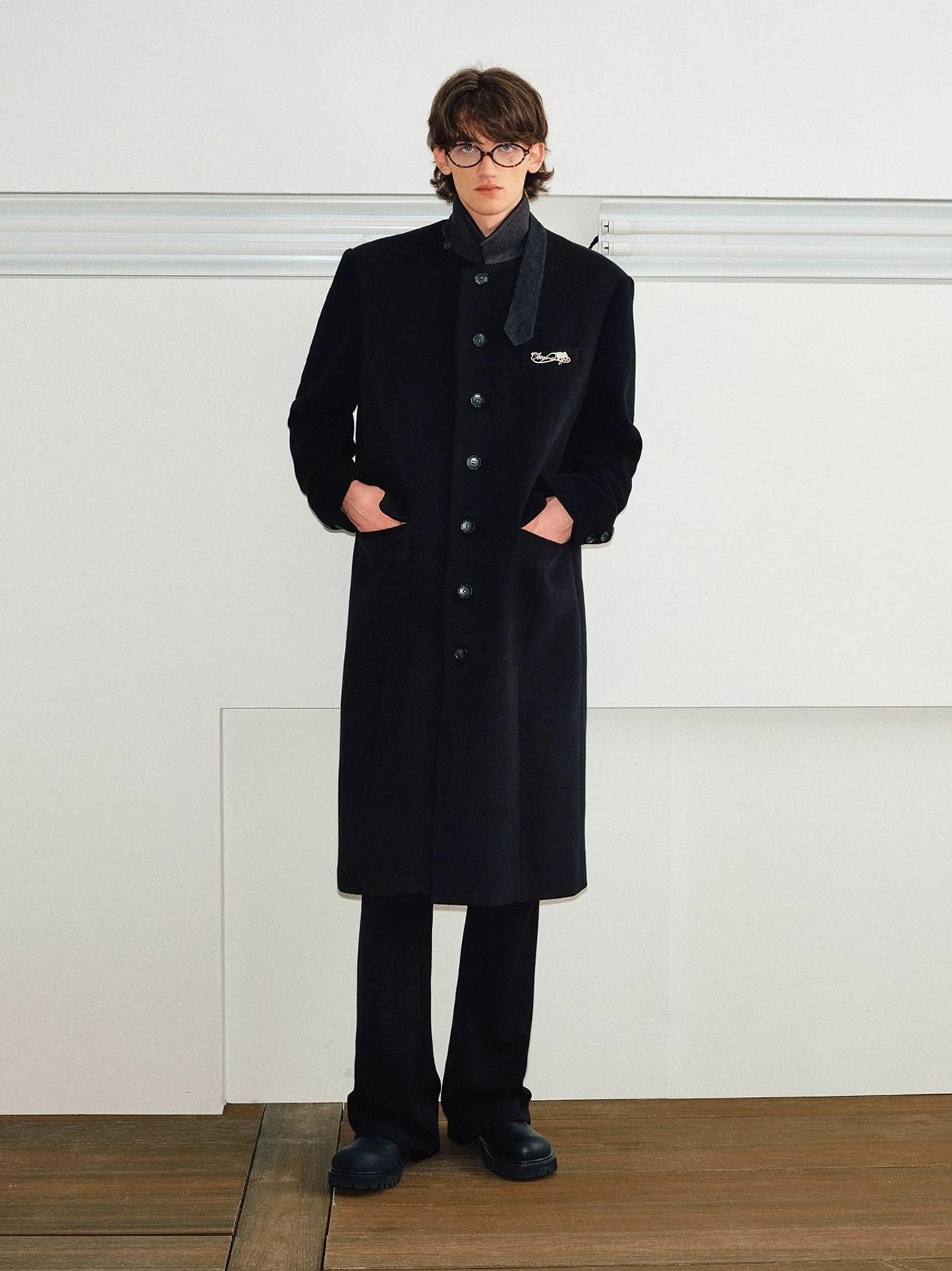 Black long wool blend stand collar overcoat worn with hands in side pockets in a minimalist studio