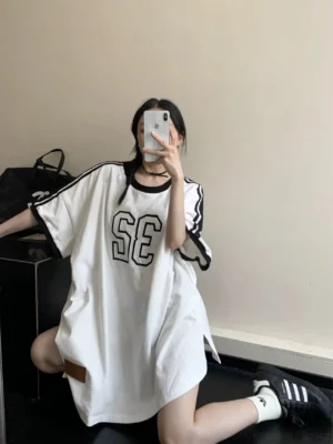 Woman wearing an oversized white varsity 32 jersey tee dress with black trim, sporty streetwear silhouette, and leather hem label in an indoor mirror selfie