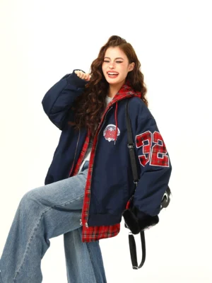Three-quarter view of a relaxed varsity layered bomber jacket with a bold number 32 sleeve patch, red plaid trim, zipper front, and black crossbody bag