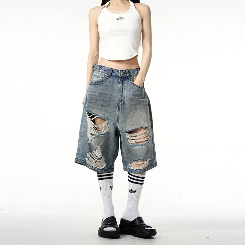Female model in a cropped white tank top styling vintage blue oversized ripped denim Bermuda shorts with loafers and striped socks