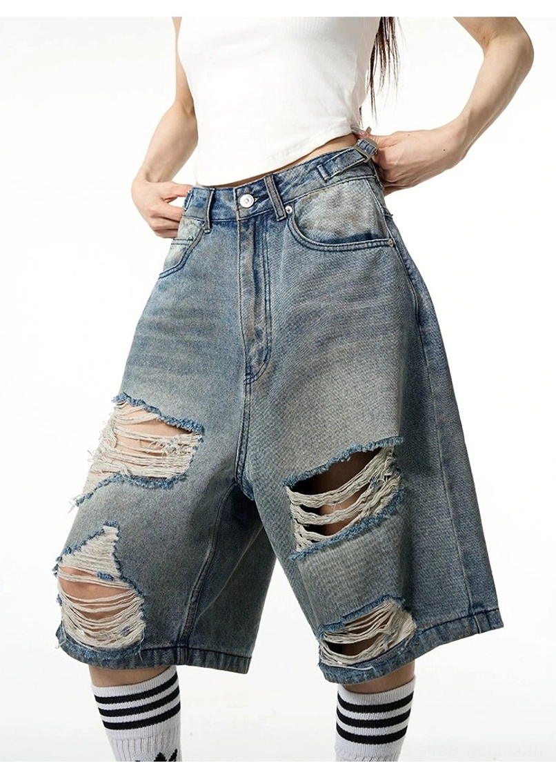 Side front view of female model wearing high-waisted looking baggy blue distressed ripped denim Bermuda shorts