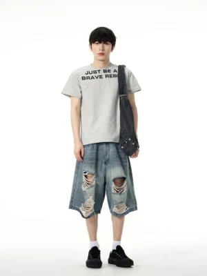 Model styling baggy vintage blue ripped denim Bermuda shorts with a graphic T-shirt and crossbody bag in a full front view