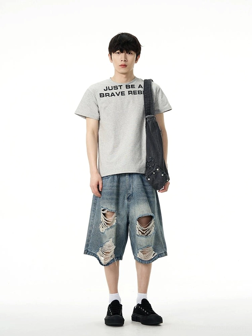 Model styling baggy vintage blue ripped denim Bermuda shorts with a graphic T-shirt and crossbody bag in a full front view