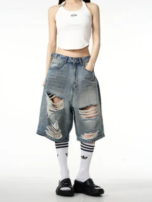 Female model wearing vintage blue distressed ripped oversized denim Bermuda shorts with a white camisole in a studio front view
