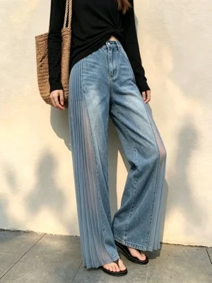 Three-quarter front view of washed blue wide-leg denim jeans featuring a pleated sheer side panel and classic zip fly
