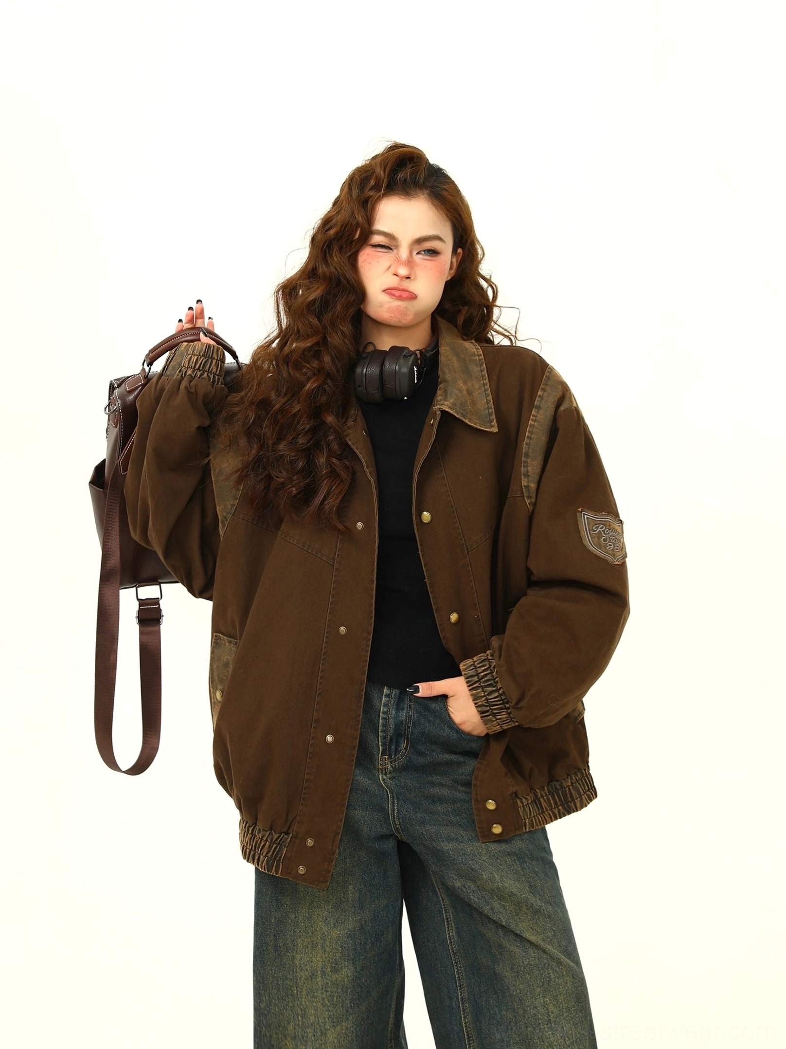 Front full-body view of a woman wearing a vintage brown oversized varsity jacket with embroidered sleeve patches, contrast shoulder panels, snap buttons, wide-leg jeans, and a brown backpack