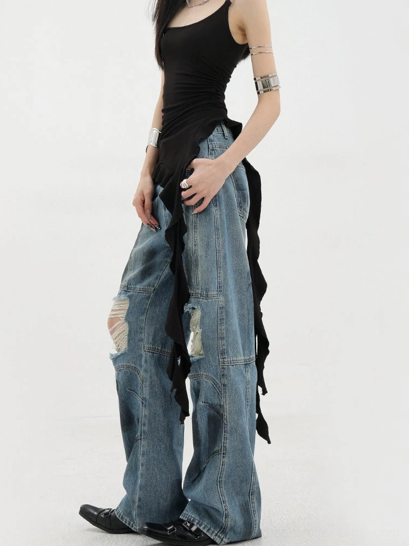 Vintage distressed wide-leg jeans left three-quarter side view on female model wearing black asymmetric camisole in studio setting showcasing faded blue wash ripped knee details loose silhouette