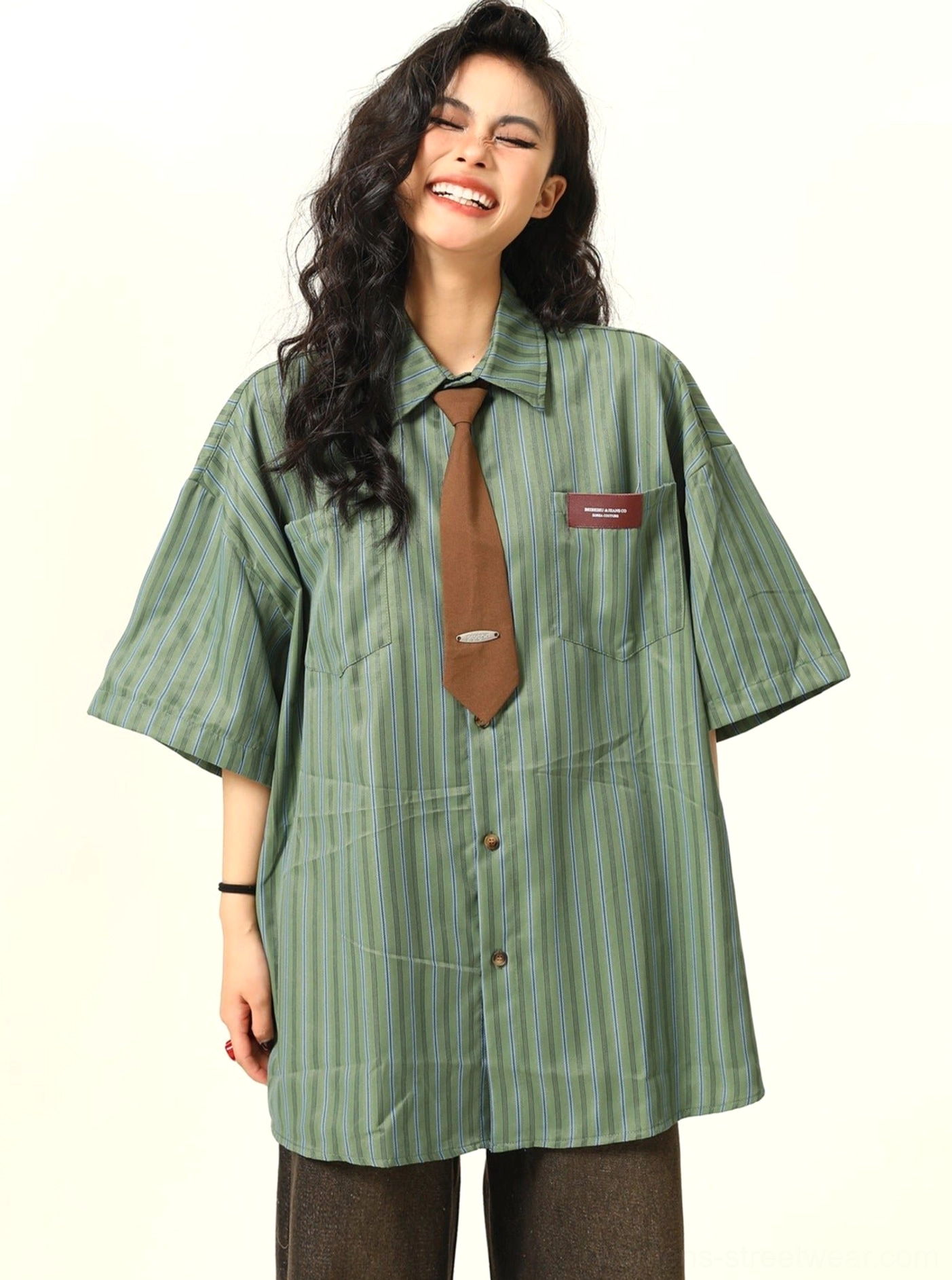 Female model wearing a vintage green striped oversized short sleeve shirt with coordinating brown tie, dark brown wide leg pants, and leather shoes in a studio setting