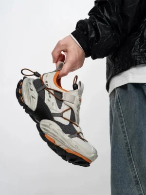 Grey orange and black tech trail sneakers shown from a low three quarter angle with breathable mesh panels, crossed webbing straps, chunky rugged outsole, and orange sole accents styled with jeans and a black streetwear jacket