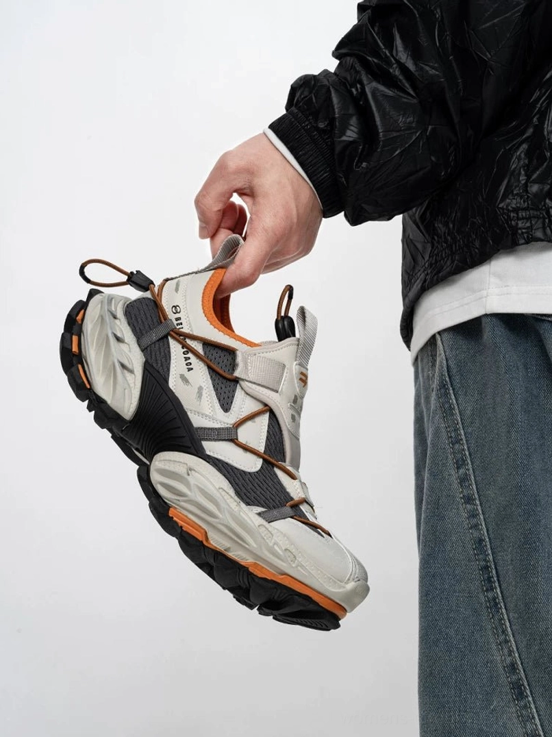 Grey orange and black tech trail sneakers shown from a low three quarter angle with breathable mesh panels, crossed webbing straps, chunky rugged outsole, and orange sole accents styled with jeans and a black streetwear jacket