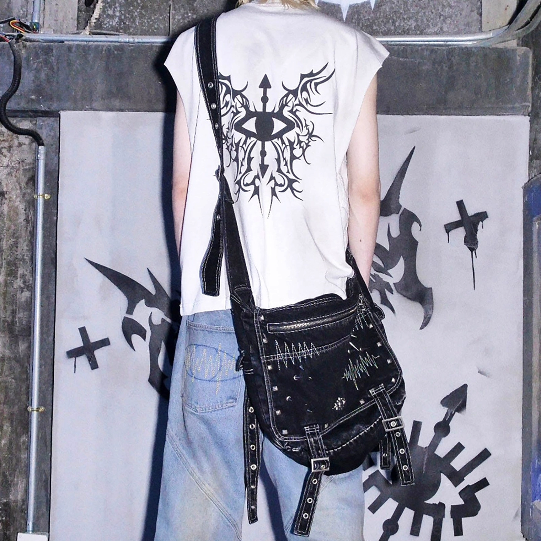 Back view of model wearing washed black denim cyberpunk crossbody bag with wide adjustable shoulder strap in industrial punk setting