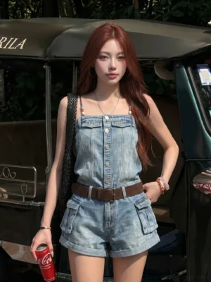 Front view of a women's washed blue beaded denim short romper with slim straps, chest pockets, rolled cargo shorts, and brown belt styling