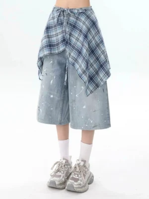 Front low angle view of washed blue distressed denim culottes with plaid overlay waist detail, white paint splatter accents, white crew socks, and chunky gray sneakers