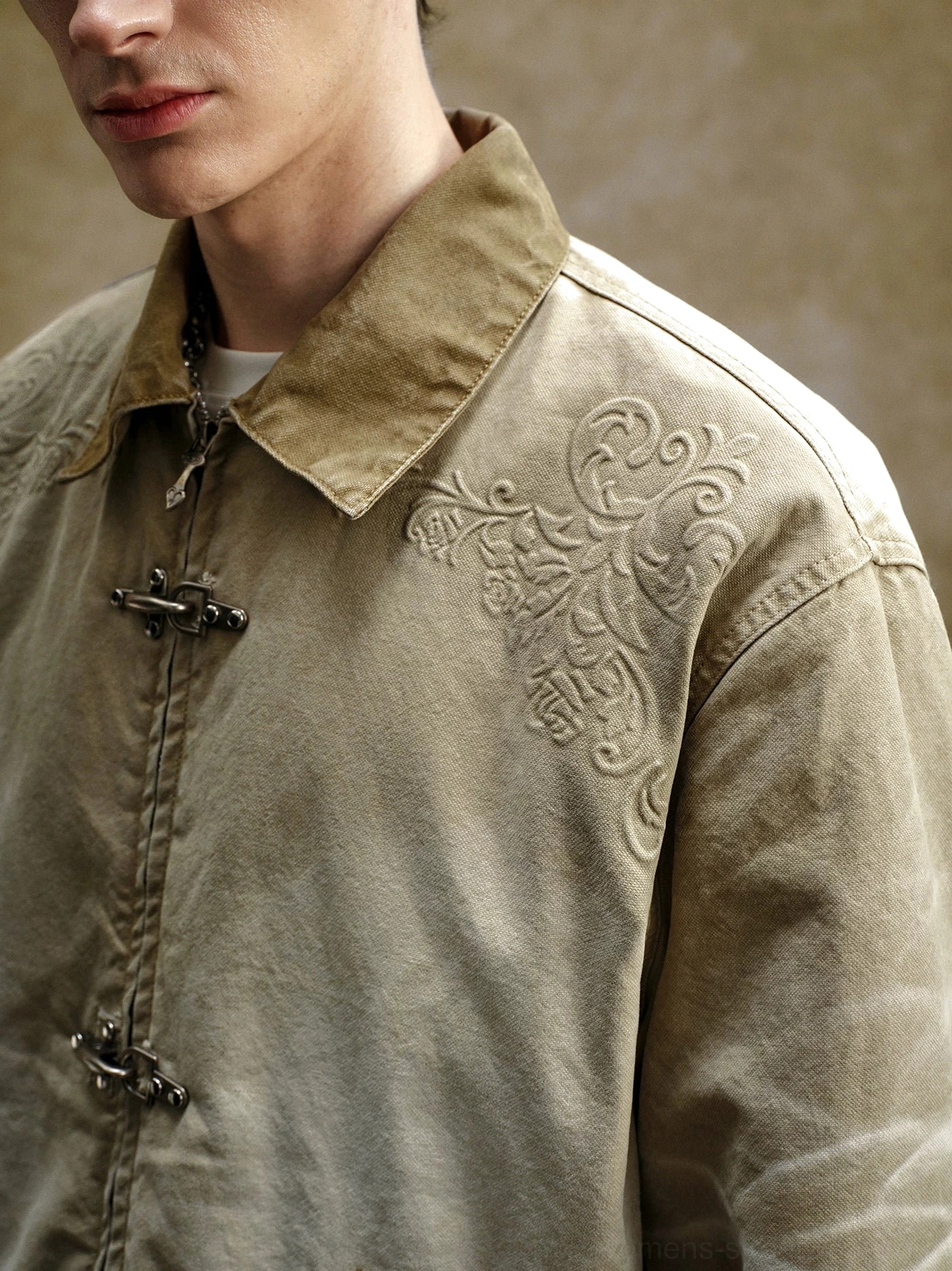 Close-up view of washed khaki embroidered utility jacket showing metal hook toggles, contrast collar, exposed placket, and shoulder embroidery