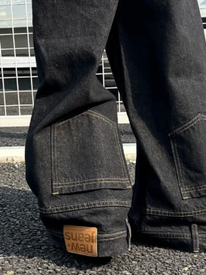 Close-up of washed black wide-leg utility denim jeans on outdoor gravel showing contrast topstitching, back patch pocket details, leather-style heel tag, and textured streetwear denim