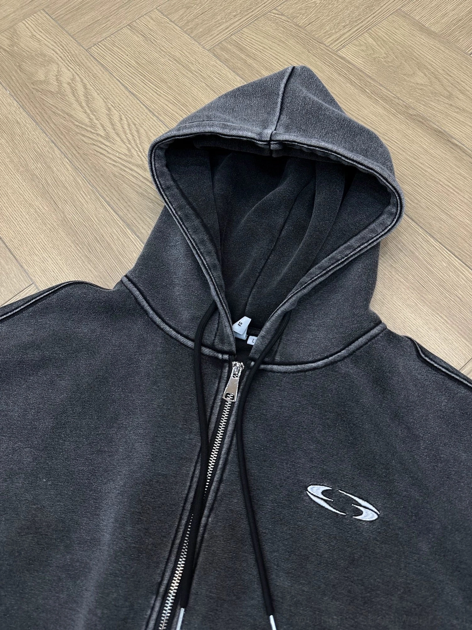Flat lay of a dark gray water-washed cropped zip-up hoodie showing the attached hood, drawstrings, full metal zipper, and chest embroidery