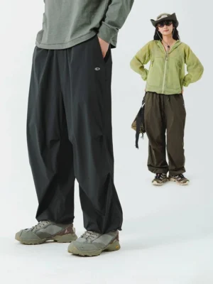 Two models wearing black wide-leg waterproof streetwear pants with an olive hooded outdoor jacket in a low-angle urban fashion scene