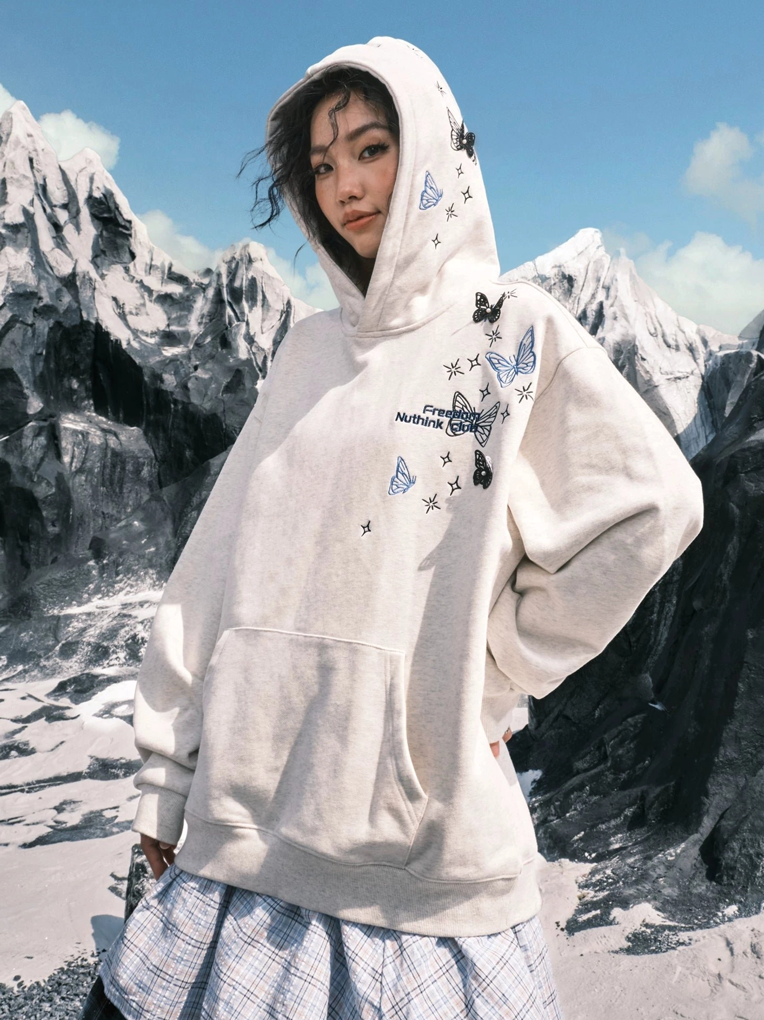 Female model wearing a White Ash butterfly embroidered oversized hoodie with hood and kangaroo pocket against a snowy mountain background