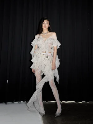 Full-body front view of a white asymmetrical lace ruffle two-piece outfit with tiered off-shoulder details, long side drape and sheer tights