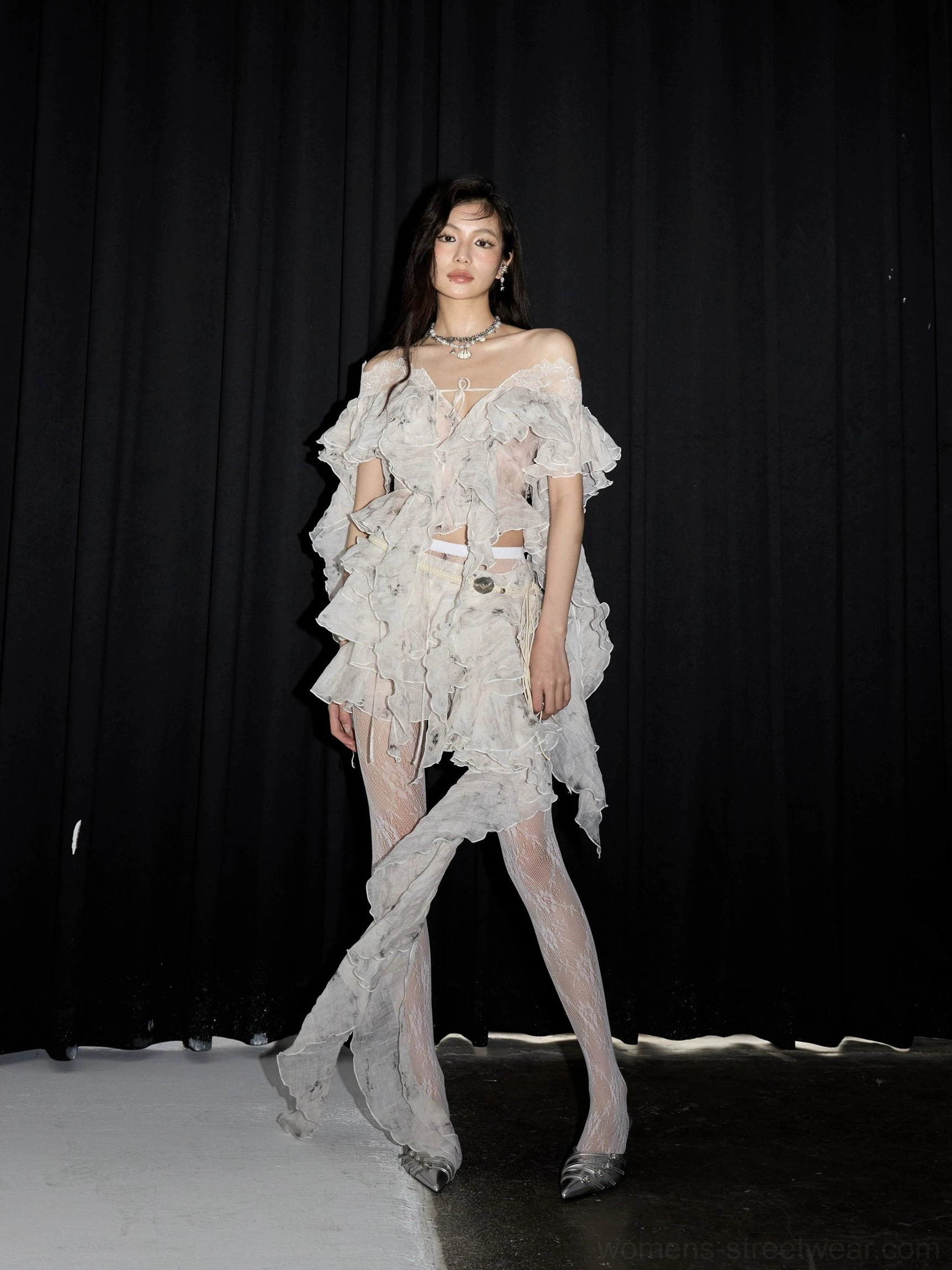 Full-body front view of a white asymmetrical lace ruffle two-piece outfit with tiered off-shoulder details, long side drape and sheer tights