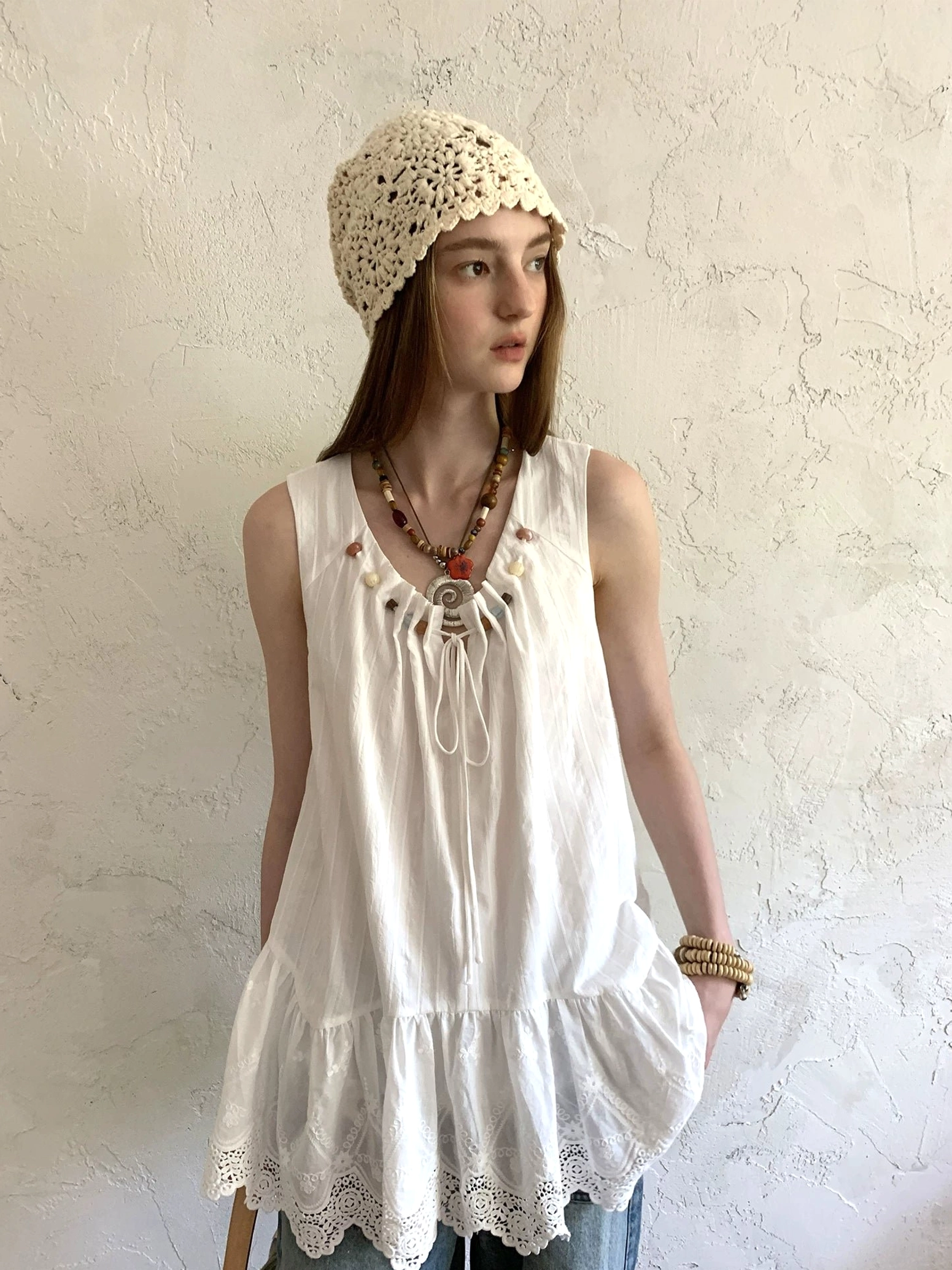 White beaded eyelet lace tunic top in a three-quarter front view, styled with blue jeans, layered jewelry, and a crochet hat